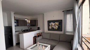 Una sala de estar moderna con un sofá gris, una mesa de centro de vidrio y una cocina al fondo.