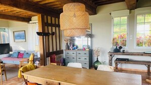 Une table à manger en bois avec des chaises, un luminaire suspendu en fibres tressées, un canapé avec des coussins, une commode et une fenêtre ornée de fleurs.