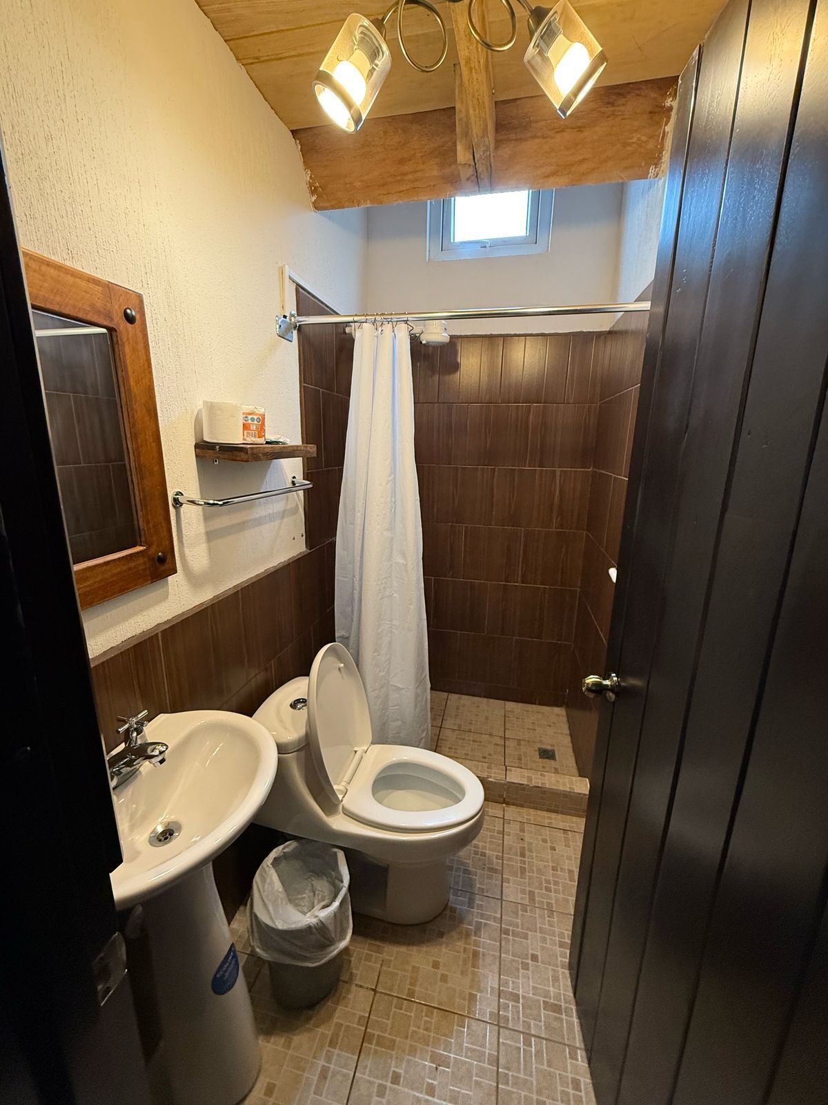 A compact bathroom with a toilet, bidet, and sink, wooden paneling, a small window, and a shower curtain.