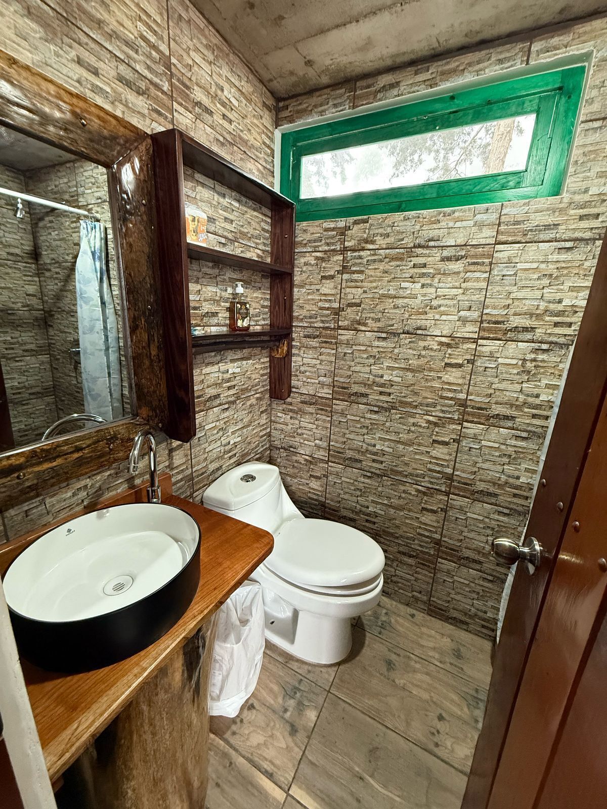 A bathroom with a stone wall design, a wooden vanity with a sink, a toilet, and a green-framed window.