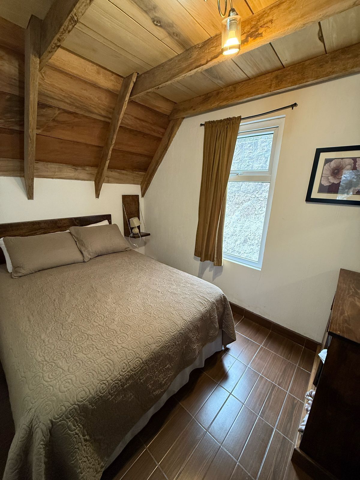 Een slaapkamer met een houten plafond, een bed met een beddeken met dessin, een raam met gordijnen en een ingelijst schilderij aan de muur.