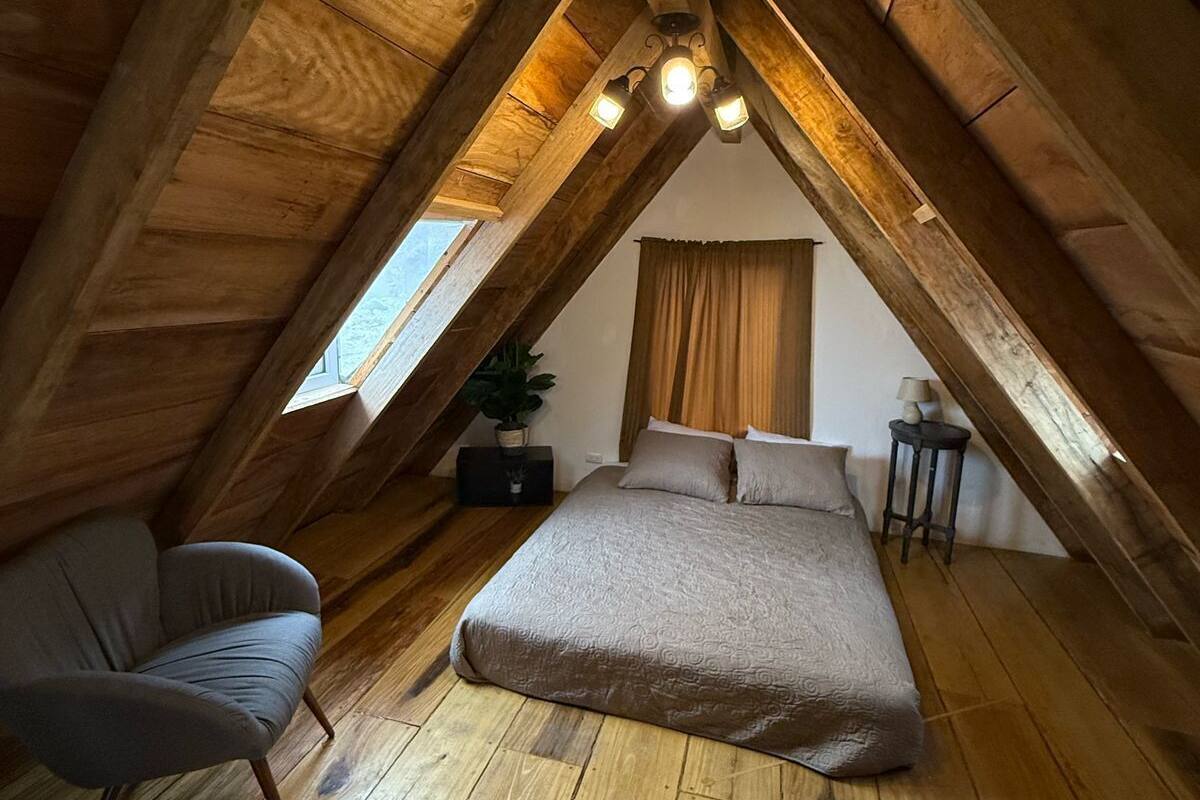 A cozy attic bedroom with a wooden sloped ceiling, a bed, a chair, and a small table.