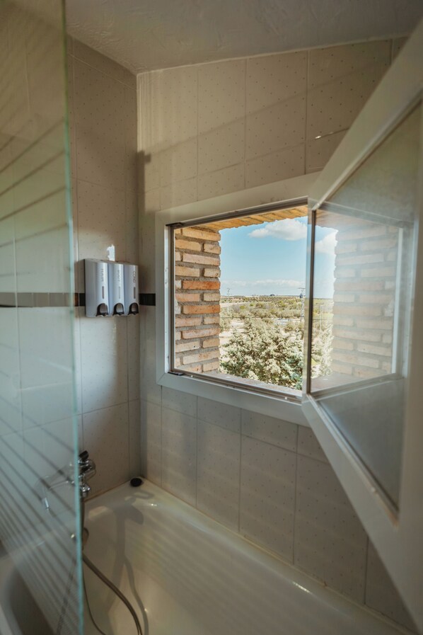 A modern bathroom with a glass shower enclosure, a bathtub, and a window with a view of a brick wall and greenery.