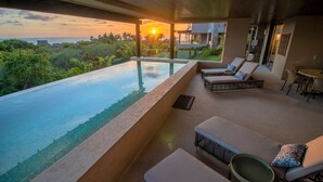 A modern infinity pool with a view of the ocean at sunset, lounge chairs, and a small table.