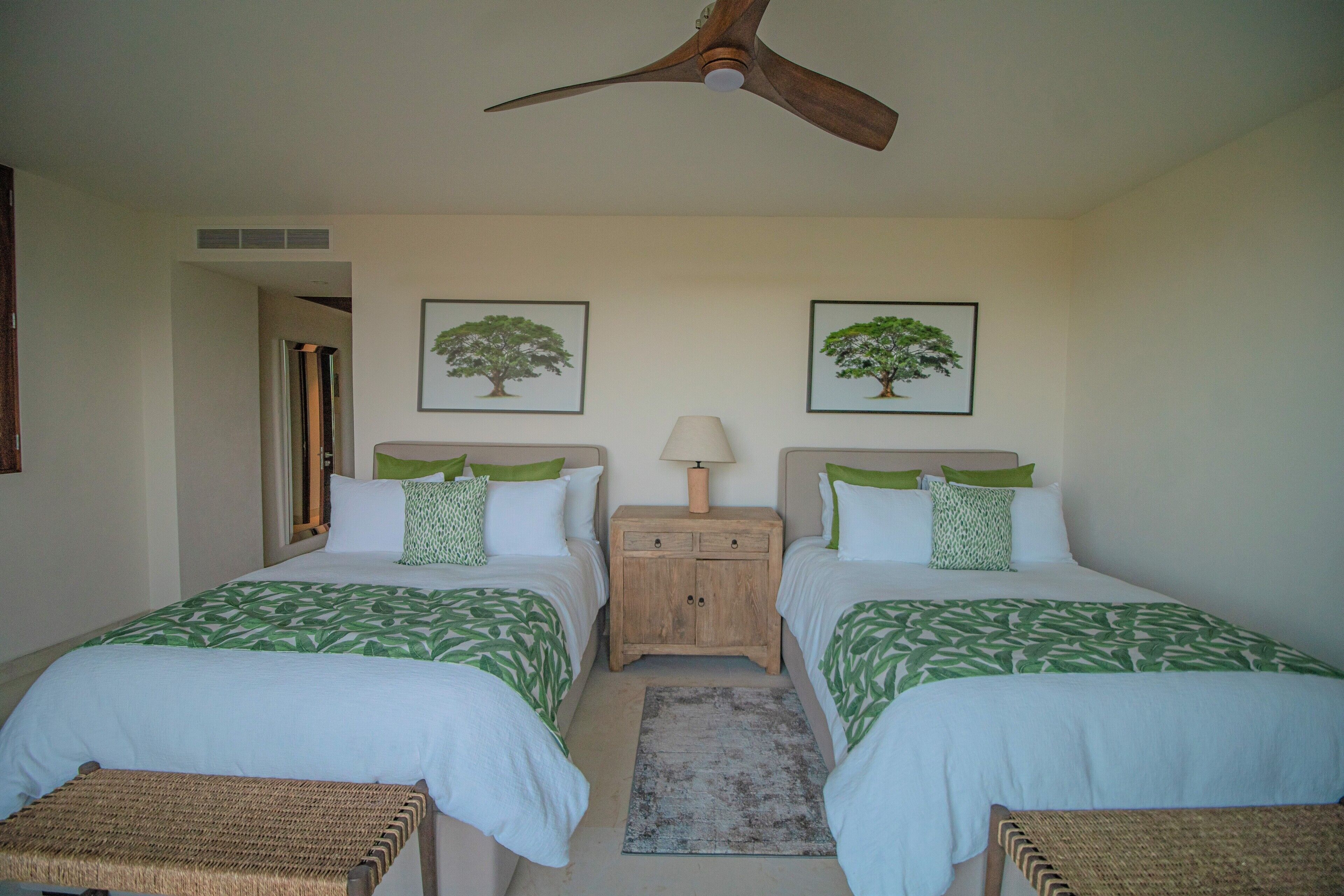 A bedroom with two beds, a wooden nightstand, a ceiling fan, and two framed pictures of trees.