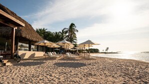 A beach with lounge chairs and umbrellas, a thatched-roof building, and palm trees.