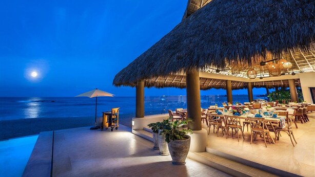 A beachfront restaurant with a thatched roof, outdoor seating, and a view of the ocean at night.
