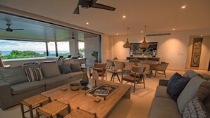 A spacious living area with a large sofa, a wooden coffee table, and a dining table. There is a view of the outdoors through large windows.