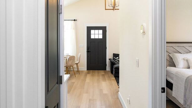 A narrow hallway with a black door, a dining area with chairs, and a bed with a headboard.