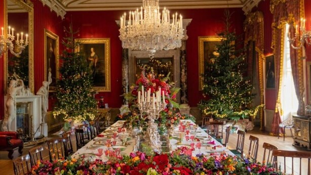 A grand dining room with a long table set for a meal, flanked by two Christmas trees and a large chandelier.