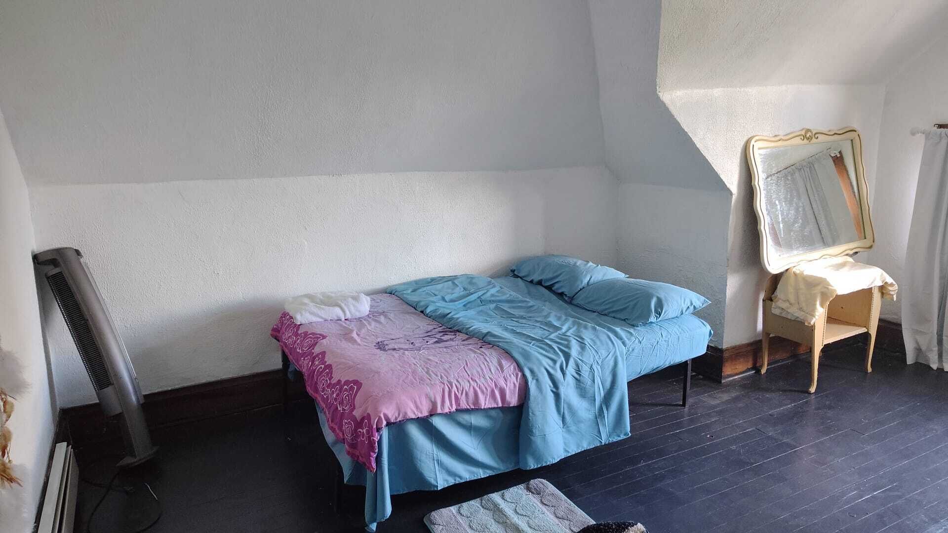 Ein kleines Zimmer mit einem Einzelbett, schrägem Dach, einem Heizlüfter, einem auf Ständer stehenden Spiegel und einem dunklen Holzboden.