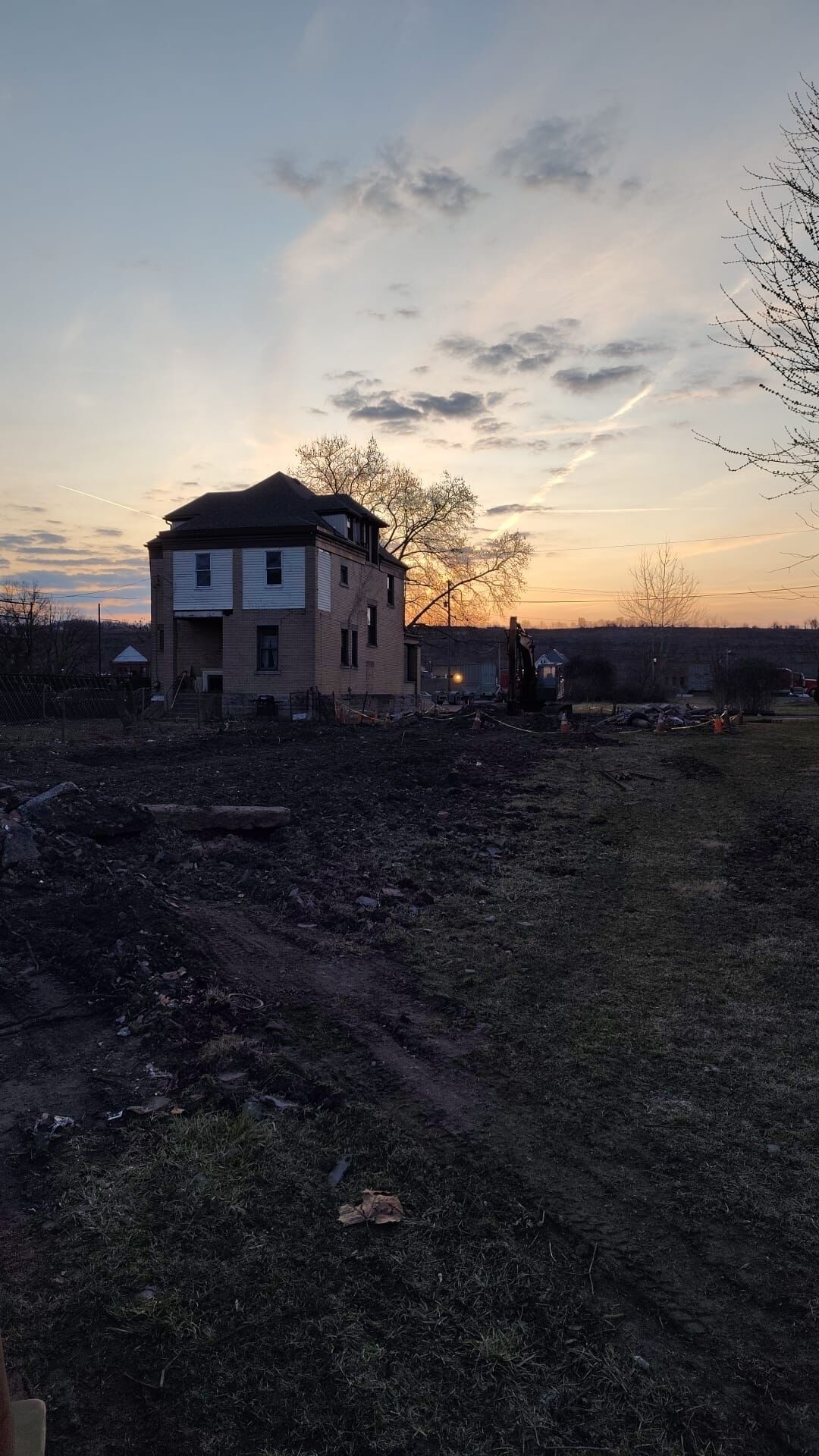 Een gebouw met twee verdiepingen, met een bakstenen gevel en een plat dak, omgeven door een vrijgemaakte plek met wat afval en een boom op de achtergrond.
