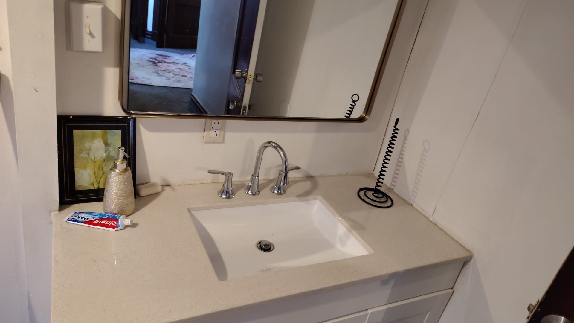 Une salle de bain avec un lavabo blanc, deux robinets, un tableau encadré, un distributeur de savon et un miroir.