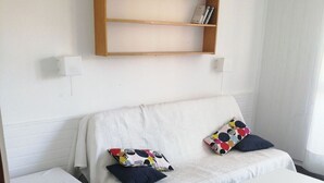 Een kamer met een witte bank, twee kussens, een klein wit tafeltje en een boekenplank met boeken en planten.