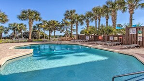 Un espace piscine avec une eau claire, des chaises longues, des palmiers et des panneaux de sécurité.