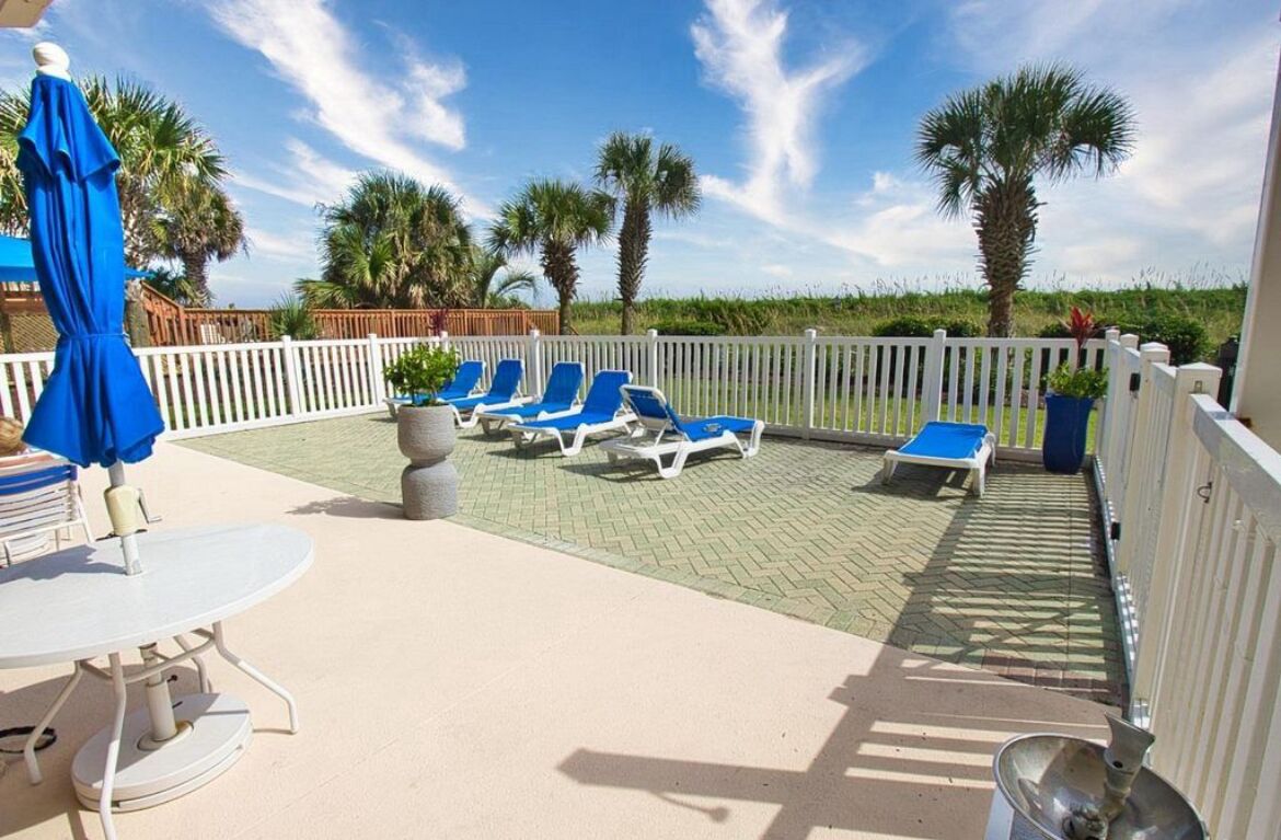 A covered outdoor area with lounge chairs and a table, surrounded by a white fence and palm trees.