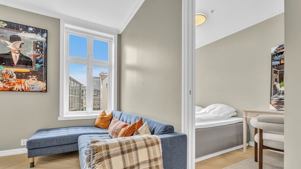 A modern living room with a blue sectional sofa, a grey bed, and a large window.