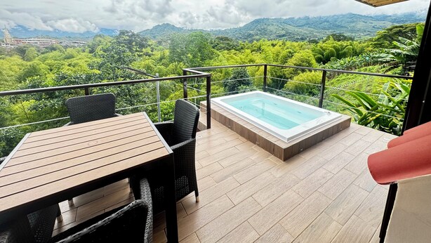 A balcony with a wooden table and chairs, a hot tub, and a view of lush green hills. - Pachakuti Ecovida Hotel (Caicedonia)