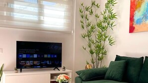 A modern living room with a flat-screen TV mounted on a white console, a green sofa, a small round table with a vase, and a colorful abstract painting on the wall.