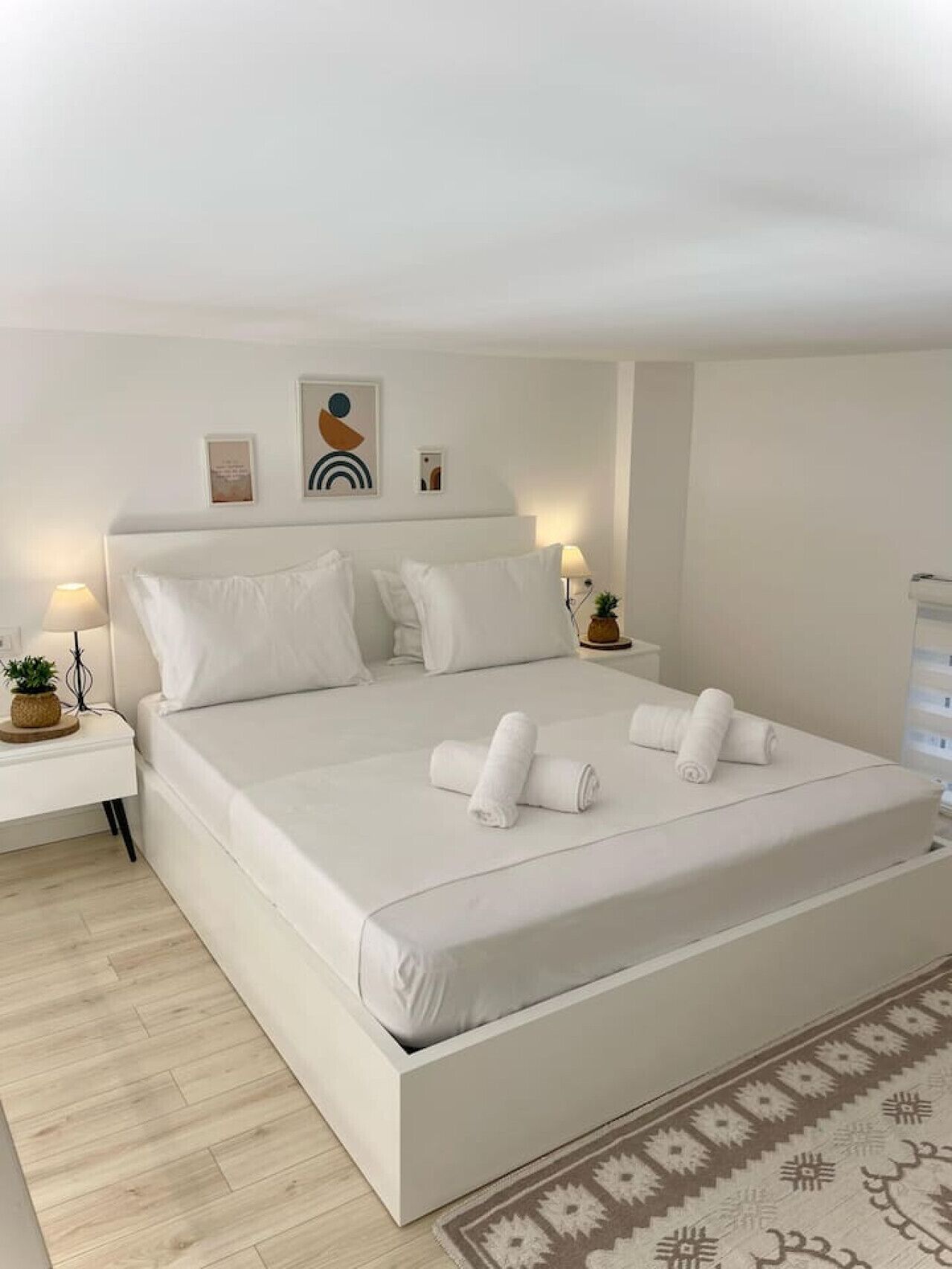 A modern bedroom with a large bed, white bedding, and a small bedside table with a lamp and a plant.