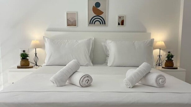 A neatly made bed with white linens and two rolled towels, flanked by two bedside tables with lamps and small potted plants.