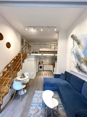 Una sala de estar moderna con un sofá azul, una zona de comedor con una mesa redonda y sillas, una cocina con lavadora, y una escalera que conduce a un nivel superior.