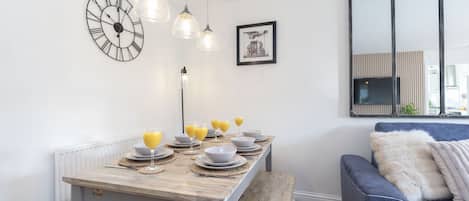 A dining area with a wooden table set for four, a clock, pendant lights, and a framed picture on the wall.