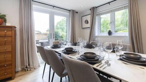 A dining area with a table set for six, large windows, and a view of greenery outside. - Stylish 5-Bed Detached Home Close to Cheltenham (Gloucestershire)