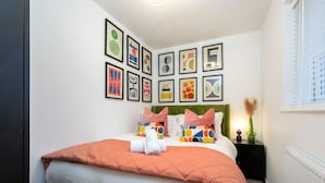 A neatly made bed with a green headboard, white and orange bedding, and colorful pillows. There are framed artworks on the wall above the bed. - Stylish 5-Bed Detached Home Close to Cheltenham (Gloucestershire)