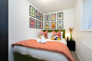 A neatly made bed with a green headboard, white and orange bedding, and colorful pillows. There are framed artworks on the wall above the bed.
