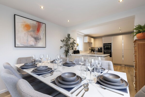 A modern dining area with a long table set for six, surrounded by grey upholstered chairs.