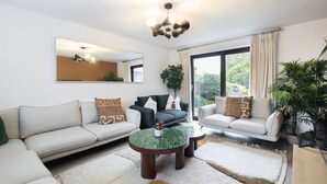 A modern living room with a large mirror, two sofas, a coffee table, and a view of greenery through the window. - Stylish 5-Bed Detached Home Close to Cheltenham (Gloucestershire)