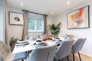 A dining area with a table set for six, including plates, bowls, and cutlery. There are two framed artworks on the walls, a window with curtains, and a potted plant on the table.
