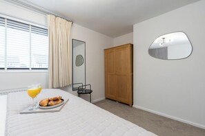 A bedroom with a bed, a bedside table with a tray of croissants and orange juice, a wooden wardrobe, a mirror, and a window with blinds.