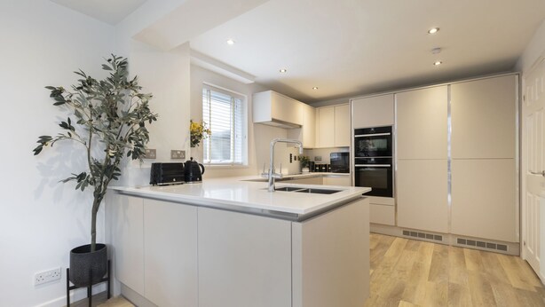 A modern kitchen with white cabinetry, a central island, a built-in oven, and a window with blinds. - Stylish 5-Bed Detached Home Close to Cheltenham (Gloucestershire)