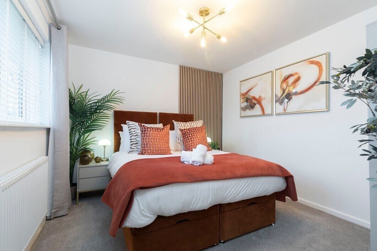 A modern bedroom with a large bed, a bedside table with a lamp, a radiator, and a window with blinds.