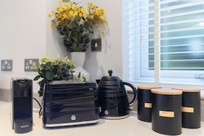 A kitchen counter with a KRUPS coffee maker, a black electric kettle, a white vase with yellow flowers, and three black containers labeled "TRICOLOR" and "SUGAR".