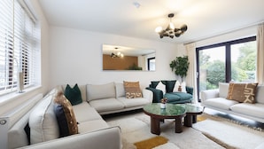 A modern living room with a sectional sofa, a coffee table, and a large window with curtains. - Stylish 5-Bed Detached Home Close to Cheltenham (Gloucestershire)