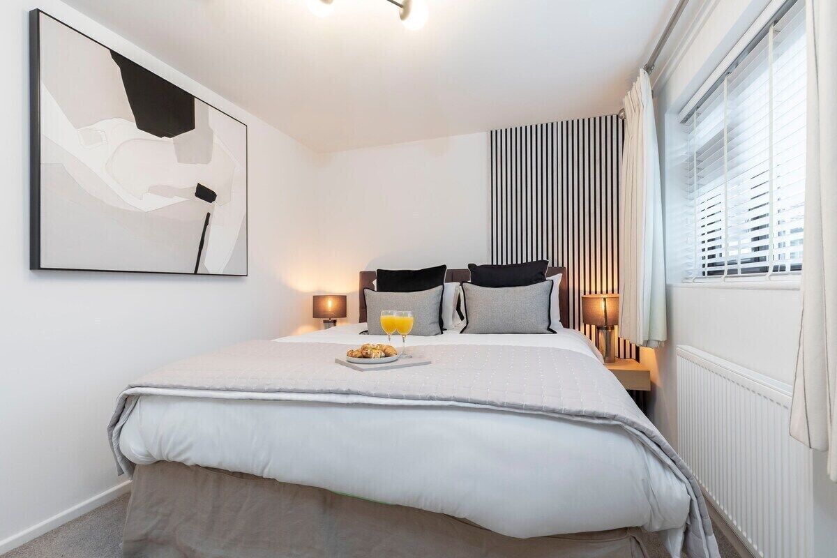 A modern hotel room with a neatly made bed, a bedside table with a lamp, a window with blinds, and a wall-mounted abstract painting.