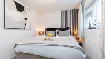 A modern hotel room with a neatly made bed, a bedside table with a lamp, a window with blinds, and a wall-mounted abstract painting.