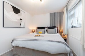 A modern hotel room with a neatly made bed, a bedside table with a lamp, a window with blinds, and a wall-mounted abstract painting.