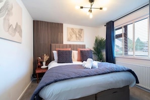A modern bedroom with a large bed, a bedside table, a nightstand, a wall-mounted artwork, and a window with blinds.