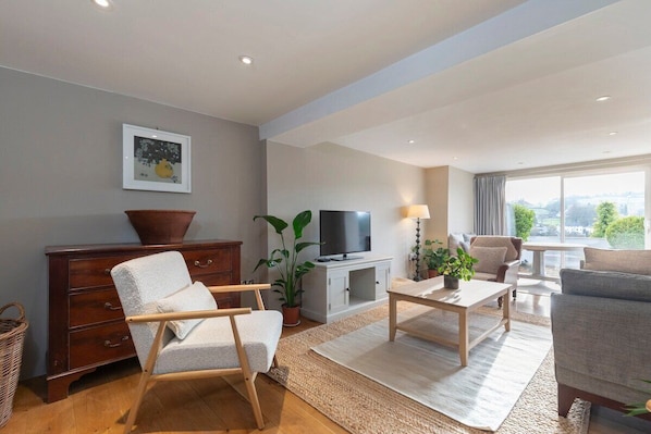 A modern living room with a sofa, armchair, TV, and a large window offering a city view. - Cotswold Hillside Retreat with Stunning Views (Stroud)