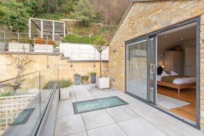 A modern bedroom with a large sliding glass door, a bed, and a view of the surrounding garden. - Cotswold Hillside Retreat with Stunning Views (Stroud)