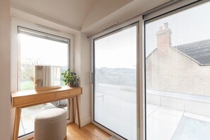 A room with a wooden desk, a cushioned stool, and a sliding glass door leading to a balcony.