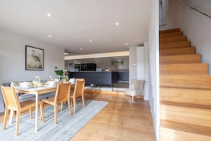 Een moderne keuken met donkere kastjes, een eethoek met houten stoelen, en een trap naar een verdieping erboven.