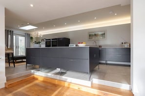 A modern kitchen with a central island, grey cabinetry, and a dining area with a chair and table. - Cotswold Hillside Retreat with Stunning Views (Stroud)
