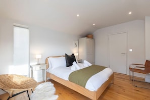 A bedroom with a wooden bed, white bedding, a wicker chair, a white fur rug, a wooden wardrobe, and a door. - Cotswold Hillside Retreat with Stunning Views (Stroud)