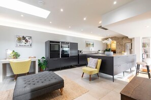 A modern kitchen with dark cabinetry, a central island, and a dining area with a grey upholstered bench. - Cotswold Hillside Retreat with Stunning Views (Stroud)
