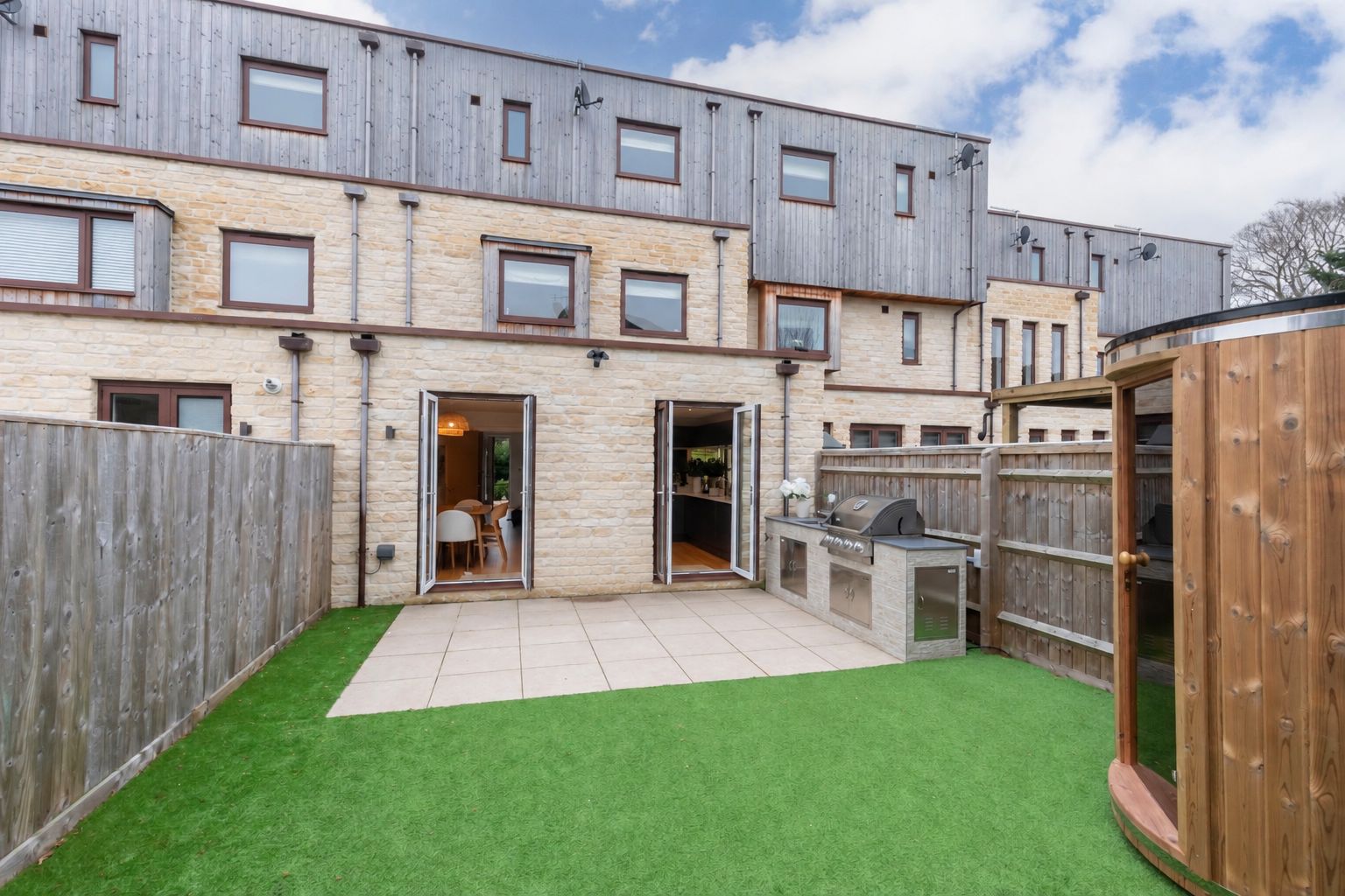 A modern two-story building with a patio area featuring artificial grass, a barbecue station, and a wooden fence.